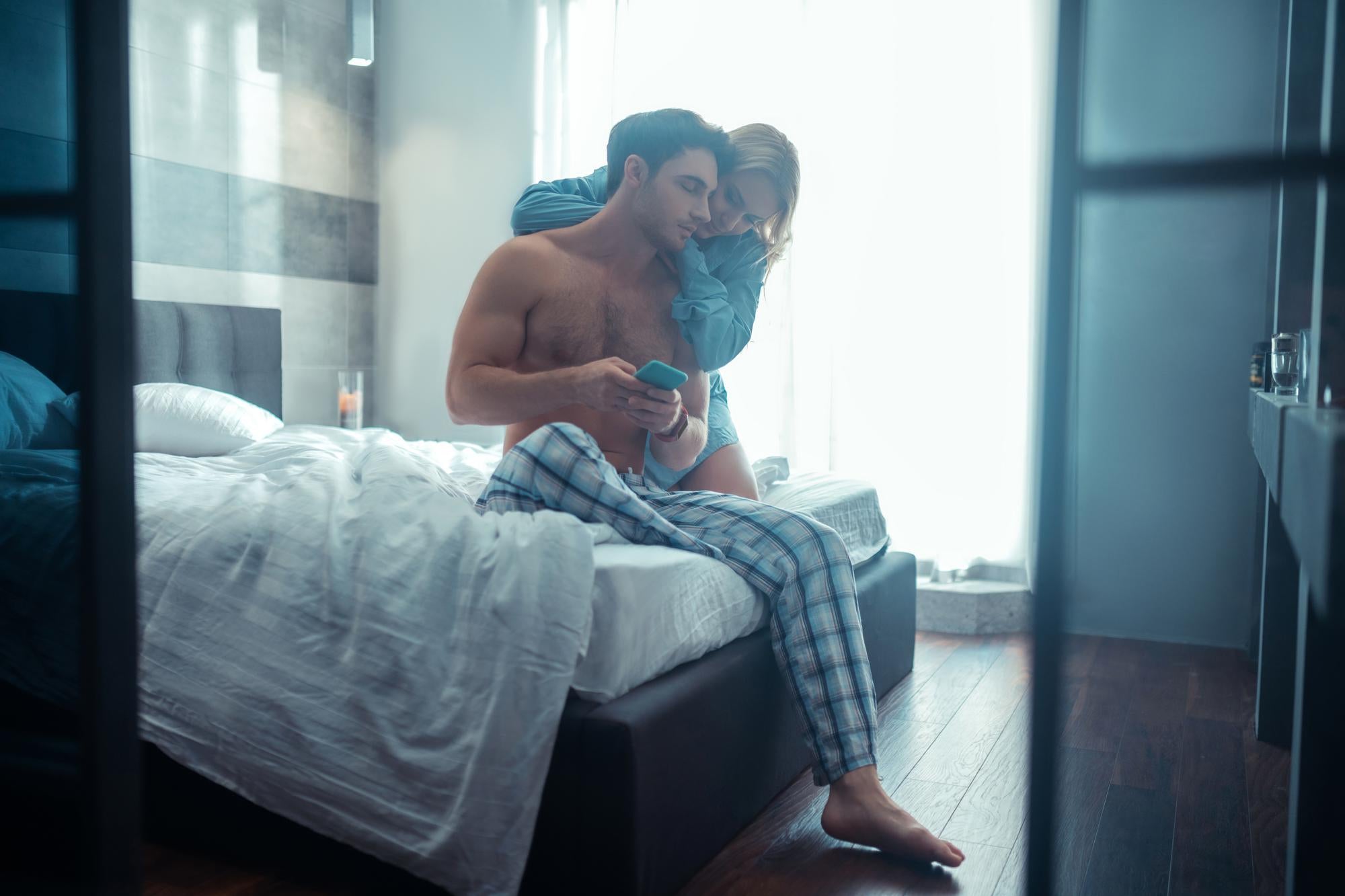 Young couple in pajamas sitting on bed with man holding a product, representing intimacy and questions about delay spray use.